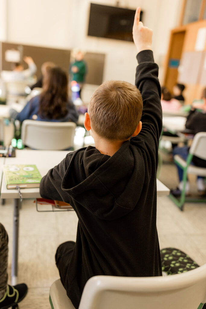 Kind sitzt im Klassenzimmer und streckt die Hand nach oben